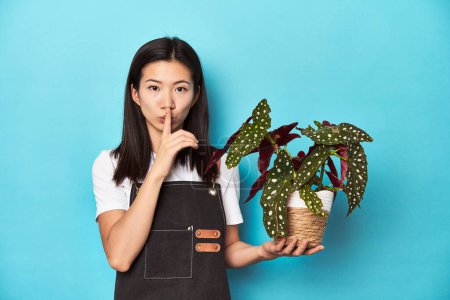 Foto de Joven jardinero asiático sosteniendo planta, estudio telón de fondo, manteniendo un secreto o pidiendo silencio. - Imagen libre de derechos