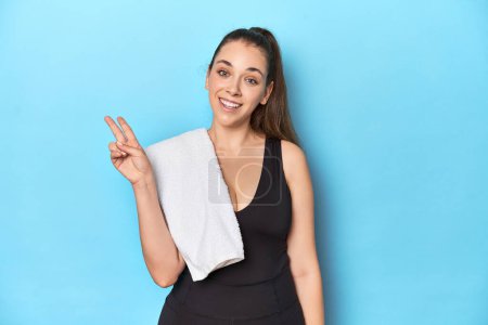 Photo for Fit young woman with a towel on her shoulder, studio shot joyful and carefree showing a peace symbol with fingers. - Royalty Free Image