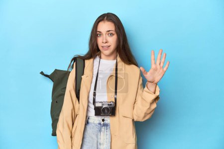 Photo for Young Caucasian woman traveler with camera and backpack smiling cheerful showing number five with fingers. - Royalty Free Image