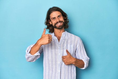 Photo for Man with beard in blue striped shirt, blue studio raising both thumbs up, smiling and confident. - Royalty Free Image