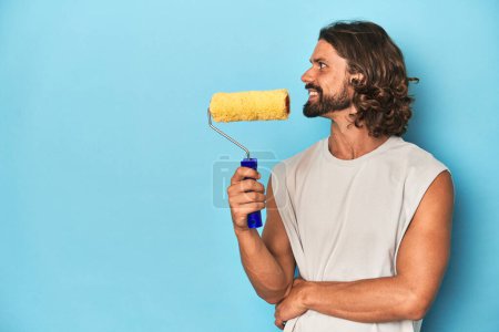 Photo for Bearded man holding a paint roller in a blue studio setting. - Royalty Free Image