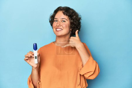 Téléchargez les photos : Jeune femme tenant un test de grossesse, fond de studio montrant un geste d'appel téléphonique mobile avec les doigts. - en image libre de droit