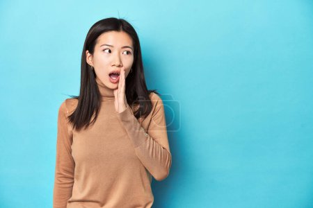 Téléchargez les photos : Jeune femme asiatique au col roulé marron, dit une nouvelle secrète de freinage chaud et regarde de côté - en image libre de droit