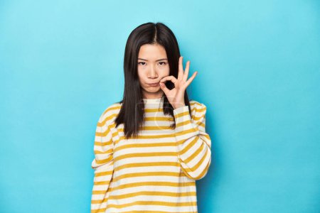 Photo for Asian woman in striped yellow sweater, with fingers on lips keeping a secret. - Royalty Free Image