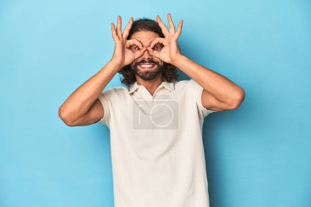 Téléchargez les photos : Homme aux cheveux longs dans un polo blanc, studio bleu montrant un signe acceptable sur les yeux - en image libre de droit
