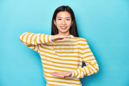 Photo for Asian woman in striped yellow sweater, holding something with both hands, product presentation. - Royalty Free Image