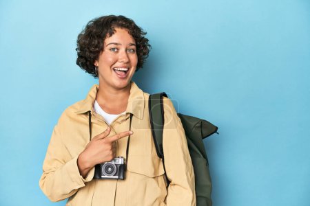 Foto de Joven viajera con cámara vintage sonriendo y señalando a un lado, mostrando algo en espacio en blanco. - Imagen libre de derechos