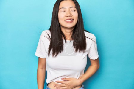 Foto de Mujer asiática joven en camiseta blanca, plano de estudio, toca la barriga, sonríe suavemente, comer y concepto de satisfacción. - Imagen libre de derechos