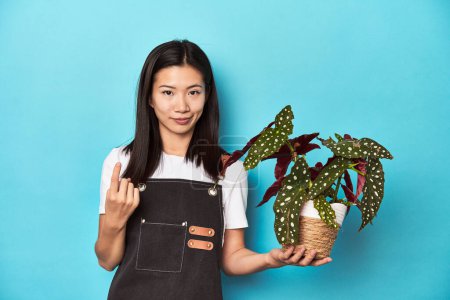 Foto de Joven jardinero asiático sosteniendo planta, estudio telón de fondo, señalando con el dedo en usted como si invitando a acercarse. - Imagen libre de derechos