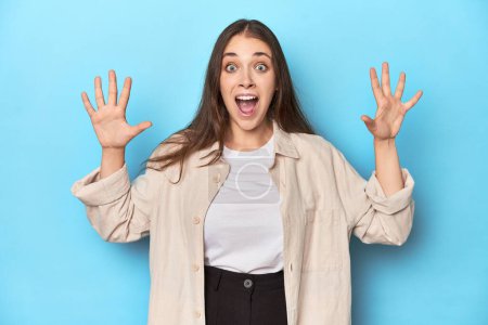 Photo for Stylish young woman in an overshirt on a blue background receiving a pleasant surprise, excited and raising hands. - Royalty Free Image