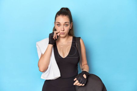 Téléchargez les photos : Jeune femme prête pour la gym avec des éléments essentiels sur fond bleu avec les doigts sur les lèvres gardant un secret. - en image libre de droit