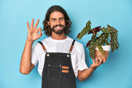 Téléchargez les photos : Jardinier aux cheveux longs avec barbe tenant une plante joyeuse et confiante montrant un geste correct. - en image libre de droit