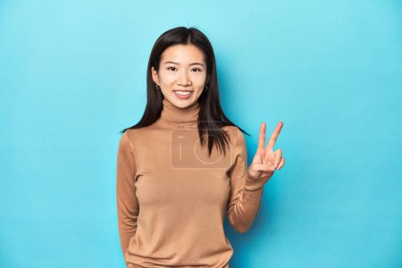 Téléchargez les photos : Jeune femme asiatique au col roulé brun, montrant le signe de la victoire et souriant largement. - en image libre de droit