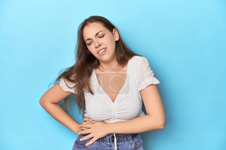 Téléchargez les photos : Jeune femme élégante en chemisier blanc sur fond de studio bleu ayant une douleur au foie, maux d'estomac. - en image libre de droit