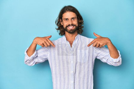 Téléchargez les photos : Homme barbe en chemise rayée bleue, studio bleu surpris pointant du doigt, souriant largement. - en image libre de droit