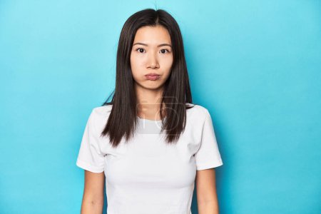 Photo for Young Asian woman in white t-shirt, studio shot, blows cheeks, has tired expression. Facial expression concept. - Royalty Free Image