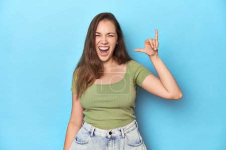 Photo for Young Caucasian woman in a green top on a blue backdrop showing a horns gesture as a revolution concept. - Royalty Free Image