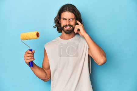 Photo for Bearded man painting with a yellow roller covering ears with hands. - Royalty Free Image
