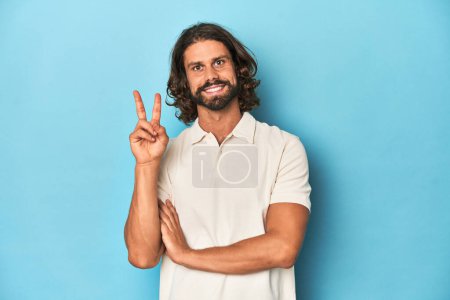 Téléchargez les photos : Homme aux cheveux longs dans un polo blanc, studio bleu montrant le numéro deux avec les doigts. - en image libre de droit
