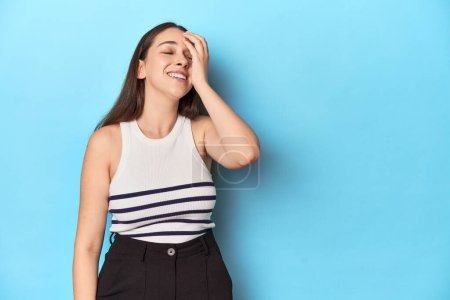 Foto de Mujer en una parte superior a rayas posando sobre un fondo de estudio azul riendo feliz, despreocupada, emoción natural. - Imagen libre de derechos