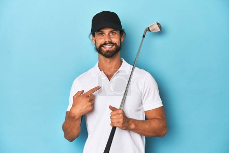 Foto de Golfista de pelo largo con club y sombrero sonriendo y señalando a un lado, mostrando algo en espacio en blanco. - Imagen libre de derechos