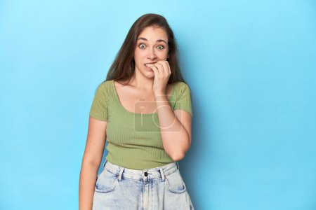Photo for Young Caucasian woman in a green top on a blue backdrop biting fingernails, nervous and very anxious. - Royalty Free Image