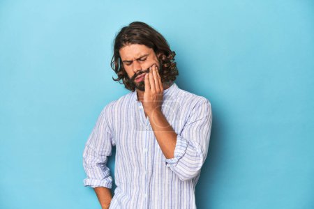 Photo for Man with beard in blue striped shirt, blue studio having a strong teeth pain, molar ache. - Royalty Free Image