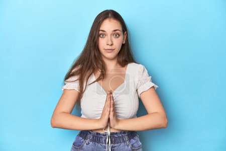 Foto de Elegante joven en blusa blanca sobre un fondo de estudio azul rezando, mostrando devoción, persona religiosa en busca de inspiración divina. - Imagen libre de derechos