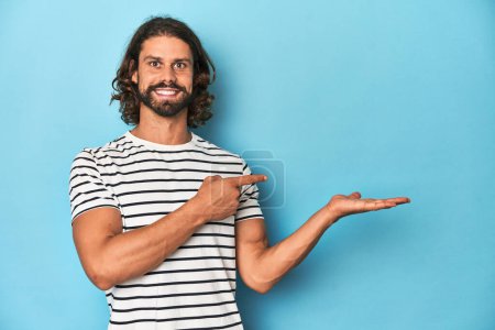 Photo for Bearded man in a striped shirt, blue backdrop excited holding a copy space on palm. - Royalty Free Image