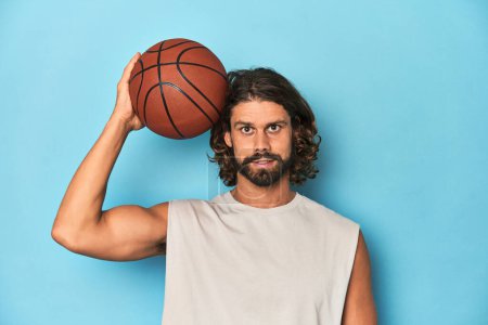 Téléchargez les photos : Homme barbu aux cheveux longs tenant un ballon de basket dans un studio bleu. - en image libre de droit