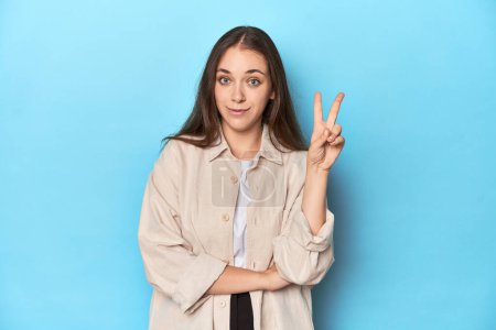 Téléchargez les photos : Jeune femme élégante dans un survêtement sur un fond bleu montrant numéro deux avec les doigts. - en image libre de droit