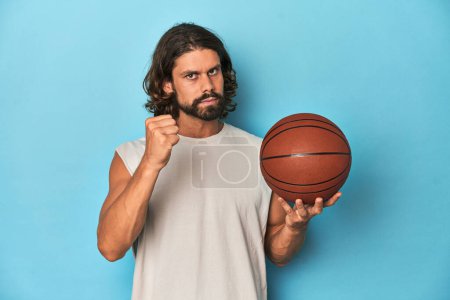 Téléchargez les photos : Homme barbu avec basket en studio bleu montrant poing à caméra, expression faciale agressive. - en image libre de droit