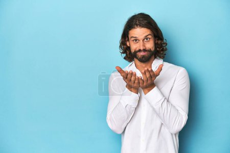 Photo for Bearded man in a white shirt, blue backdrop folding lips and holding palms to send air kiss. - Royalty Free Image