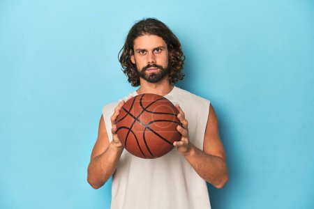 Photo for Long-haired, bearded man holding a basketball in a blue studio. - Royalty Free Image