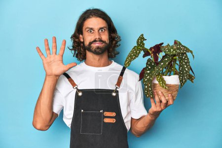 Foto de Jardinero de pelo largo con barba sosteniendo una planta sonriente alegre mostrando el número cinco con los dedos. - Imagen libre de derechos