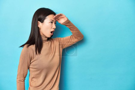Photo for Young Asian woman in brown turtleneck, looking far away keeping hand on forehead. - Royalty Free Image