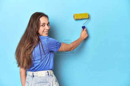 Foto de Joven mujer caucásica con rodillo de pintura amarillo en estudio azul. - Imagen libre de derechos