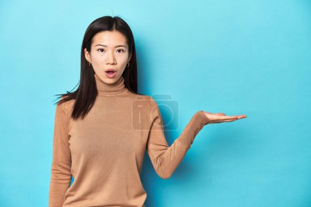 Photo for Young Asian woman in brown turtleneck, impressed holding copy space on palm. - Royalty Free Image