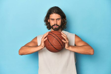 Téléchargez les photos : Homme barbu aux cheveux longs tenant un ballon de basket dans un studio bleu. - en image libre de droit