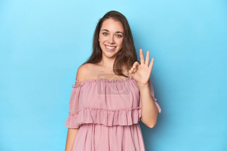 Foto de Mujer joven con un vestido rosa en un fondo de estudio azul alegre y confiado mostrando gesto ok. - Imagen libre de derechos