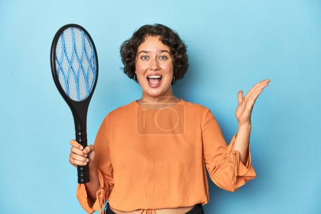 Photo for Young woman with electric mosquito racket Young woman with electric mosquito racketreceiving a pleasant surprise, excited and raising hands. - Royalty Free Image