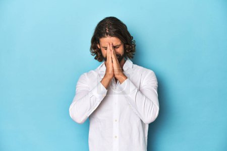 Foto de Hombre barbudo con una camisa blanca, fondo azul cogido de la mano en orar cerca de la boca, se siente seguro. - Imagen libre de derechos