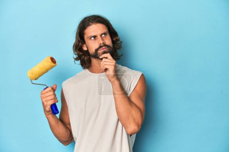Photo for Bearded man painting with a yellow roller looking sideways with doubtful and skeptical expression. - Royalty Free Image