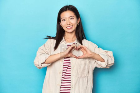 Téléchargez les photos : Femme asiatique en chemise à couches et t-shirt rayé, souriant et montrant une forme de coeur avec les mains. - en image libre de droit