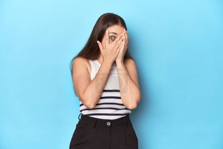 Téléchargez les photos : Femme dans un haut rayé posant sur un fond de studio bleu cligner des yeux à travers les doigts effrayé et nerveux. - en image libre de droit