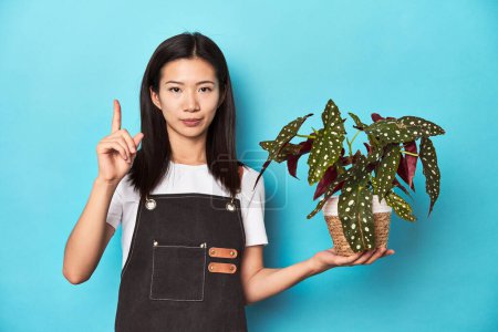Téléchargez les photos : Jeune asiatique jardinier tenant plante, studio toile de fond, montrant numéro un avec doigt. - en image libre de droit