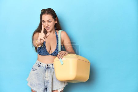 Photo for Woman in bikini with a portable beach cooler, blue studio showing number one with finger. - Royalty Free Image