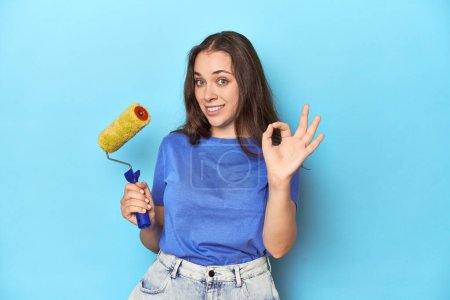 Photo for Young woman with yellow paint roller on a blue background cheerful and confident showing ok gesture. - Royalty Free Image
