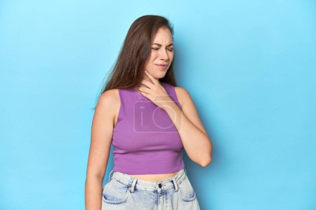 Téléchargez les photos : Jeune femme à la mode dans un haut violet sur fond bleu souffre de douleur à la gorge due à un virus ou une infection. - en image libre de droit