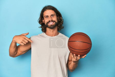 Photo for Bearded man with basketball in blue studio person pointing by hand to a shirt copy space, proud and confident - Royalty Free Image
